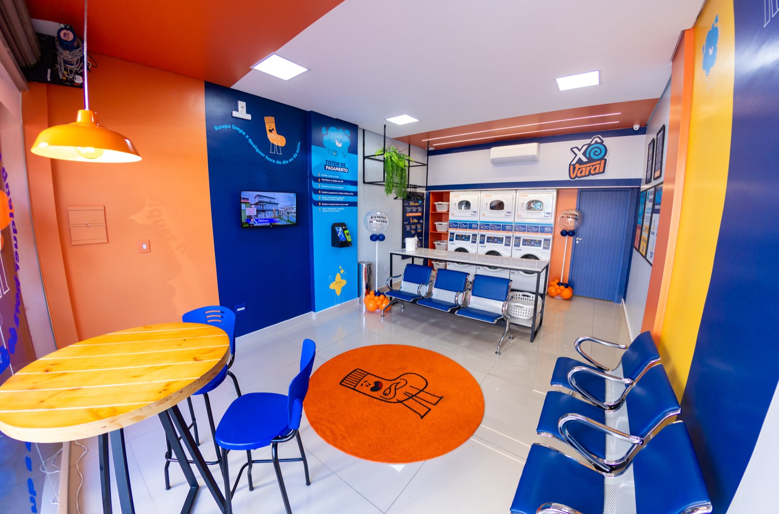 Interior de uma unidade Xô Varal: ambiente colorido em azul e laranja, com mesas de convivência e lavadoras ao fundo